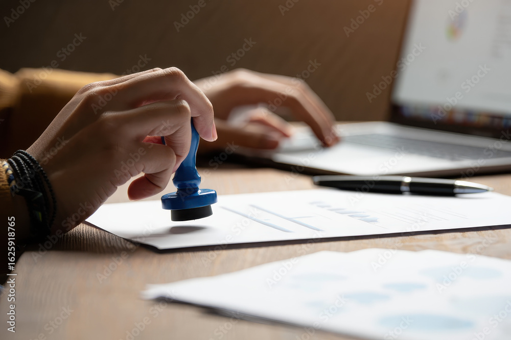 Fototapeta premium Person using a blue stamp on a document with a laptop and pen in the background on a desk