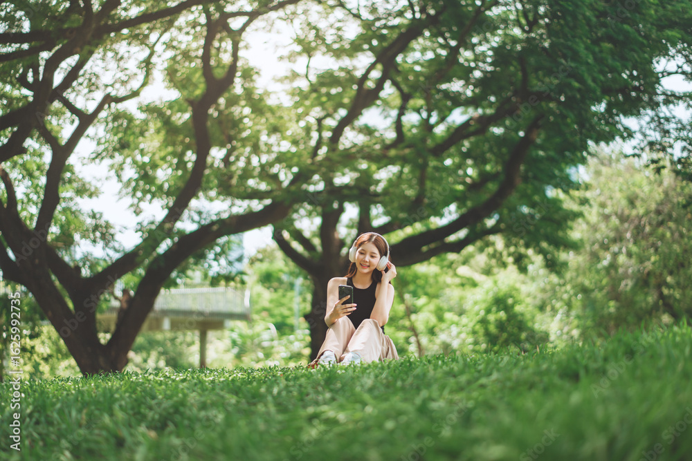 Obraz premium Cheerful asian woman wearing headphones and using smartphone to browse music or social media while sitting under a tree in city park during spring – relaxing lifestyle with soft sunlight, copy space