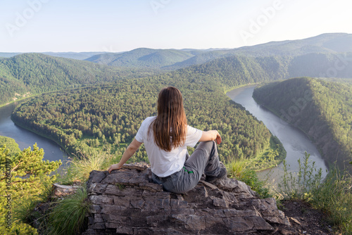 Принти на полотні A person sits on a rocky ledge, gazing at a breathtaking view of a winding river framed by rolling hills and dense forests