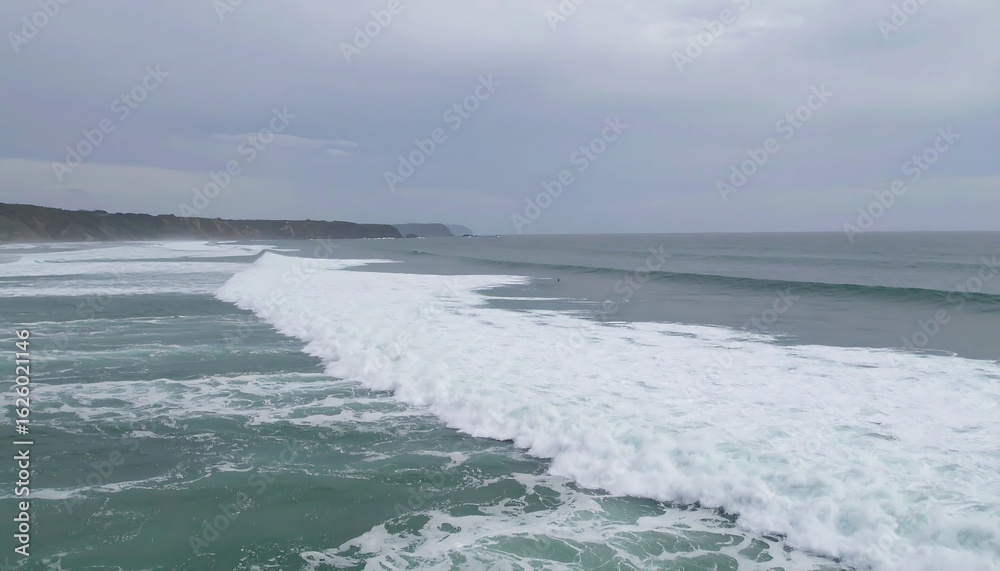 Fototapeta premium Expansive ocean view; rolling waves break near a coastline under a cloudy sky