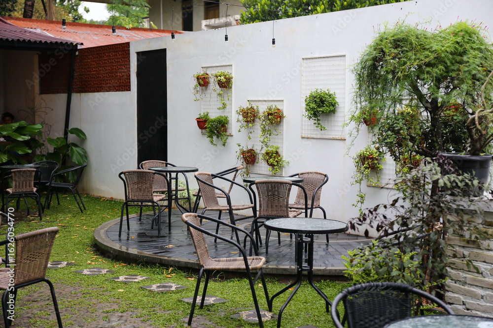 Fototapeta premium A cozy outdoor seating area of a cafe with wicker chairs and tables. Lush green plants hang on a white wall, creating a natural and inviting atmosphere perfect for relaxation.