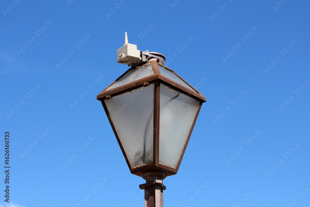Fototapeta premium An old, rusted streetlamp is topped with a small, modern wireless sensor or communication device, combining historic urban infrastructure with contemporary technology