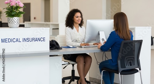 Two Women Discussing Medical Insurance at Reception Desk with White Blouse and Blue Top In Bright Office Setting with Pink Flowers and Computer