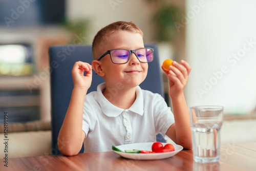 Cute boy in glasses eating vegetables at home