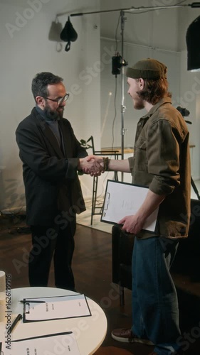 Vertical shot of young actor greeting his filming director for first time and shaking his hand in studio
