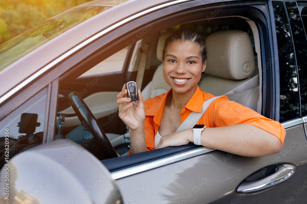 Naklejka premium Happy young black woman holding car keys from driver's seat, looking out of window. Concept of buying a new car, success, ownership, or independence, closeup