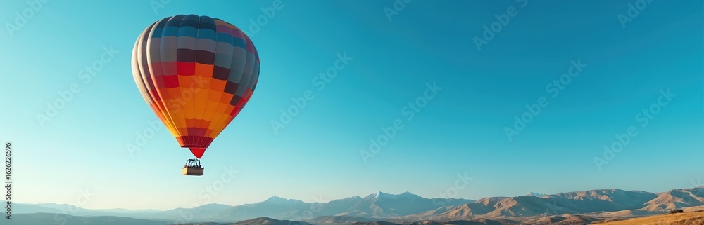 Naklejka premium Colorful hot air balloon glides across vibrant blue sky over mountainous landscape. Experience freedom, adventure, exploration. Enjoy peaceful soaring journey, breathtaking views, majestic voyage