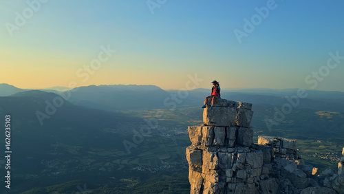 Photography A lone hiker triumphantly sits atop a rugged cliff, overlooking a vast valley in golden sunrise light
