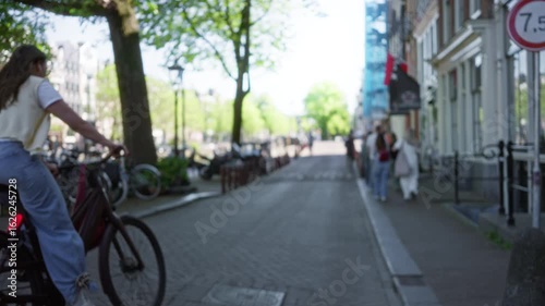 Wallpaper Mural Blurry view of a european city street with women walking and cycling past tall trees and brick buildings under a clear blue sky. Torontodigital.ca