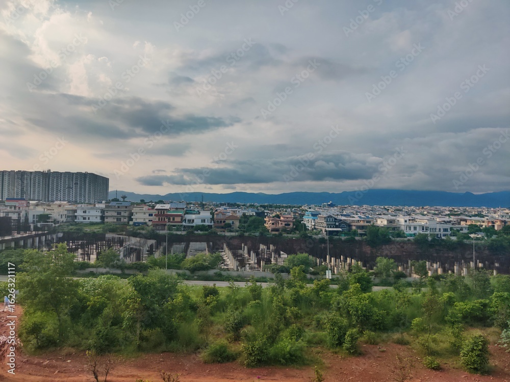 Fototapeta premium Dramatic cityscape showing urban development, a high-rise building, and lush greenery under a moody, sunlit sky.