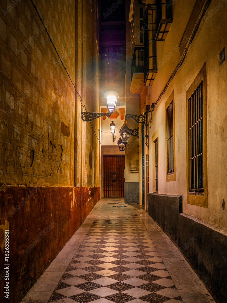 Obraz premium Tiled alley in Calle Don Carlos Alonso Chaparro, Casco Antiguo, Seville, Spain with warm walls, ornate lamps, barred windows, and a wooden door