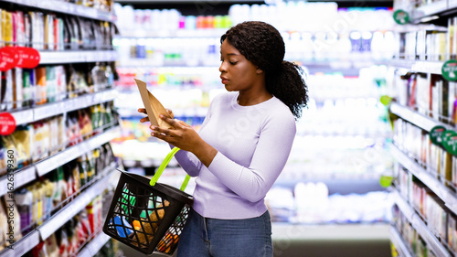 Beautiful African American woman making choice of products at supermarket. Focused female consumer shopping for groceries, selecting food, reading labels at modern mall