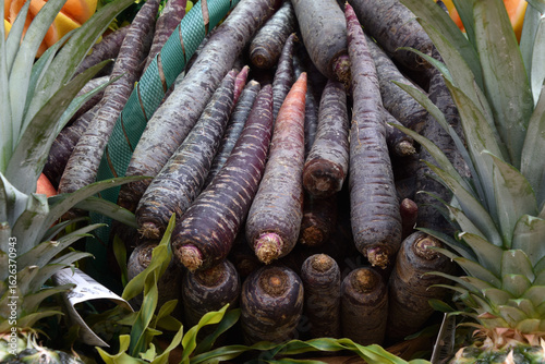 Fresh purple carrots arranged in a vibrant display with green leaves and tropical fruits, showcasing organic produce in a colorful market setting 