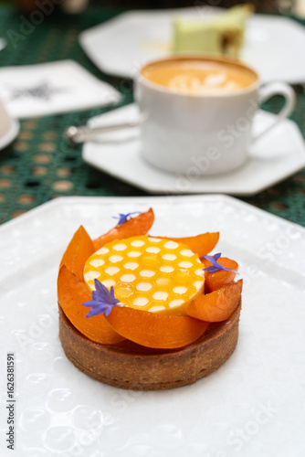 A piece of apricot tart decorated with mini purple flower on a white plate with a cup of coffee in background.