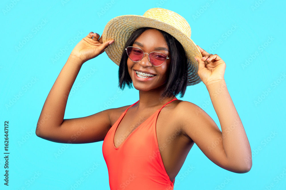 Summer fun. Portrait of cheerful African American woman posing in sexy swimsuit, wearing straw hat and sunglasses on blue studio background. Beach vacation or pool party concept Summer fun. Portrait of cheerful African American woman posing in sexy swimsuit, wearing straw hat and sunglasses on blue studio background. Beach vacation or pool party concept