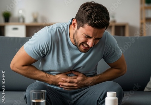 A distressed young man experiencing intense abdominal pain, clutching his stomach while sitting on the couch at home with medication nearby.