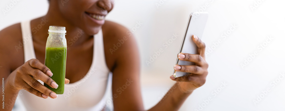 Fototapeta premium Weight Loss App. Slim Black Lady Using Phone Drinking Healthy Green Smoothie Standing In Modern Kitchen At Home. Detox Diet Recipes Application For Smartphone Concept. Shallow Depth