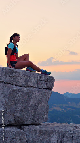 Focused female runner atop rugged cliff, dressed in vibrant gear, celebrates serene achievement against a stunning sunset. Highlights endurance, joy, and cross-country running's triumph amidst.