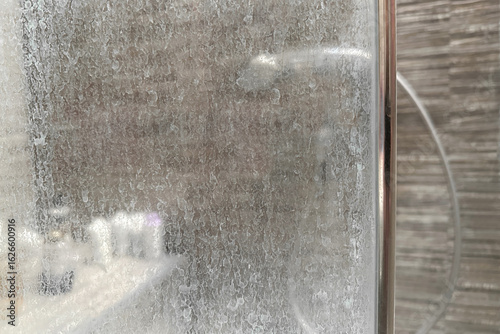 Close-up of a dirty shower glass door with white mineral residue from hard water and soap scum. Blurred shower cabin in the background. Common bathroom cleaning issue.