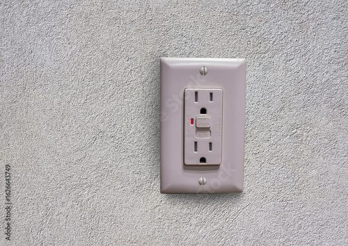 Electrical wall outlet with ground fault circuit interrupter (GFCI) showing reset and test buttons on textured white wall background.