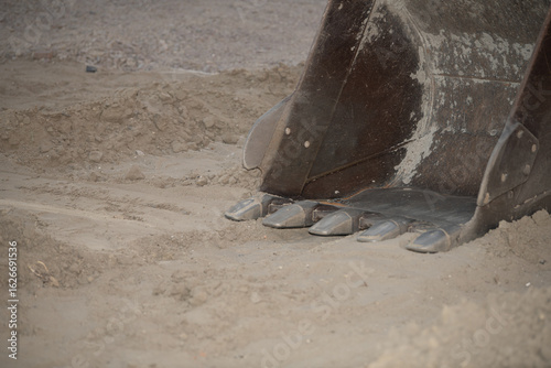 An excavator bucket rests on the sandy ground, ready to dig. The setting is a construction site during the early evening, showcasing the heavy machinery in action