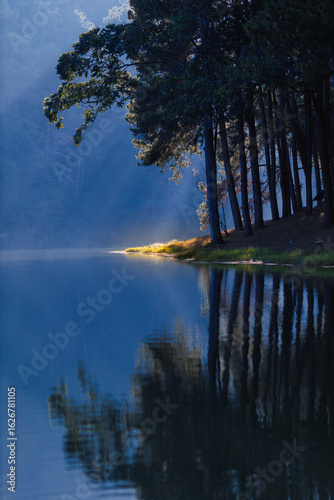 morning sunlight streaming through pine trees with mist over Pang Ung lake in Mae Hong Son, Thailand, reflecting on calm water creating a serene and peaceful atmosphere