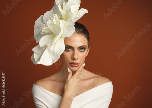 Stylish woman wearing a giant white flower headpiece, finger near lips, posing against orange background. Fashion portrait with unique floral accessory.