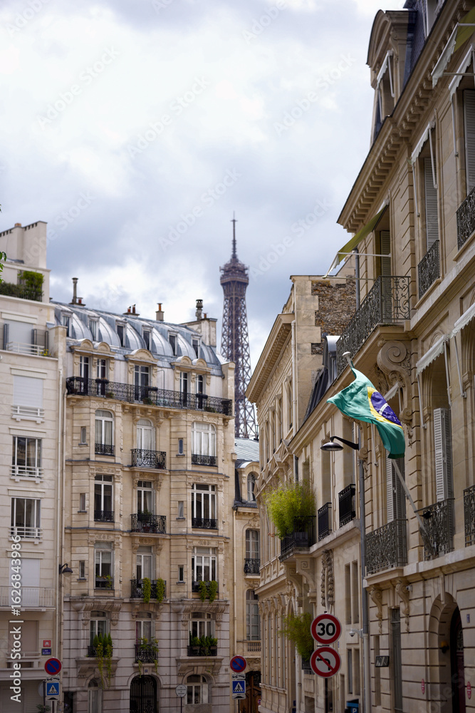 Fototapeta premium Vertical view of Eiffel Tower behind traditional Parisian buildings with French flag on cloudy day