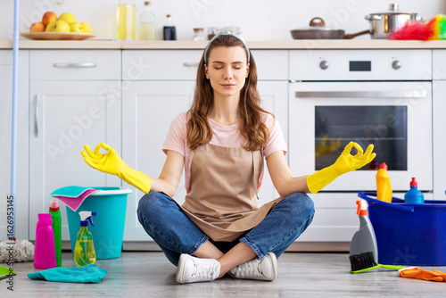 Fotografi Young housewife surrounded by cleaning supplies meditating on floor at kitchen, taking break from house chores