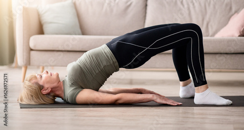 Stay at home fitness. Strong mature woman doing half bridge yoga pose, strengthening her abs muscles indoors, copy space. Fit senior woman working out in living room during covid isolation