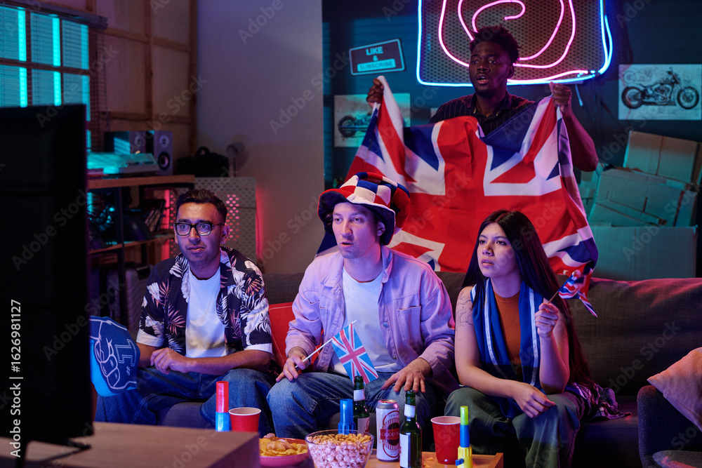 Obraz premium Group of young adults and Black man supporting British sports team, holding United Kingdom flags and watching television together, snacks and drinks on table in living room