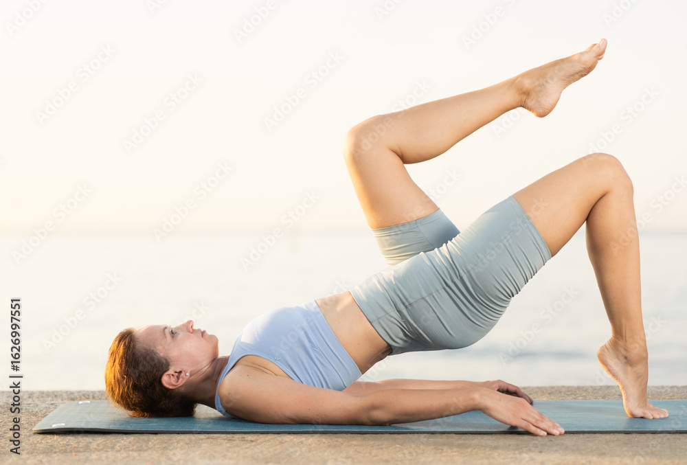 Obraz premium Sporty female wearing sportswear standing legs up in Salamba Sarvangasana exercise during yoga on seashore at sunrise outdoors