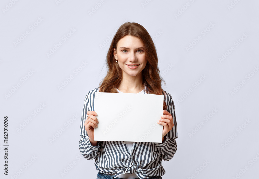 Fototapeta premium Portrait of beautiful woman holding white blank advertising board, smiling millennial lady demonstrating empty banner with copy space for your text or design, standing on gray background in studio