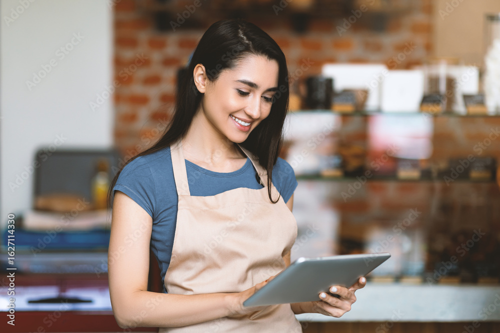 Fototapeta premium Young woman owner of modern cafe in apron holding digital tablet and standing with shopwindow on background, using pad, copy space. Modern technology and work in cafeteria