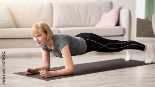 Domestic workout concept. Athletic senior woman standing in elbow plank on yoga mat at home. Sporty mature lady working out indoors, strengthening her core muscles during covid lockdown