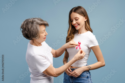 Fototapeta Senior woman attaching pink ribbon on young woman's t-shirt: breast cancer aware