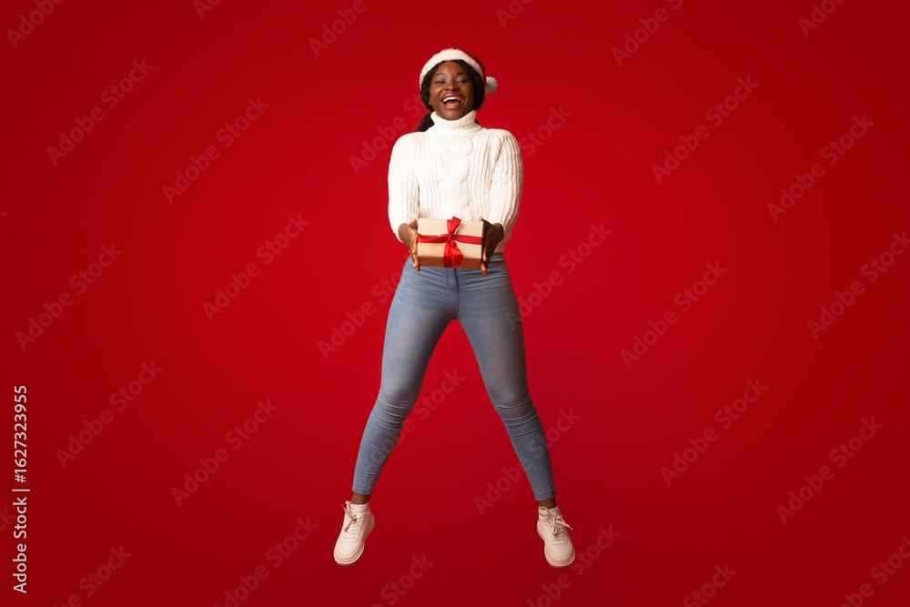 Cheerful black woman in santa hat jumping in air with christmas present in hands over orange background, excited african american lady holding wrapped present box and smiling, enjoying xmas holidays Cheerful black woman in santa hat jumping in air with christmas present in hands over orange background, excited african american lady holding wrapped present box and smiling, enjoying xmas holidays