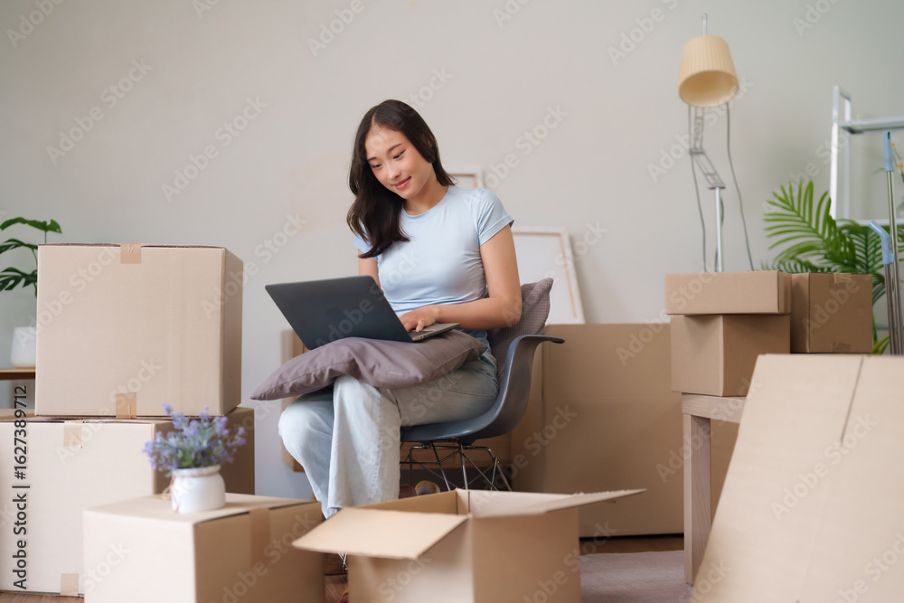 Fototapeta premium Asian adult woman actively manages new home relocation using laptop computer for planning and organizing amidst moving boxes in a modern apartment