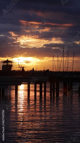 Majestic orange and purple sunset over silhouette pier as men with phones take photos in slow motion