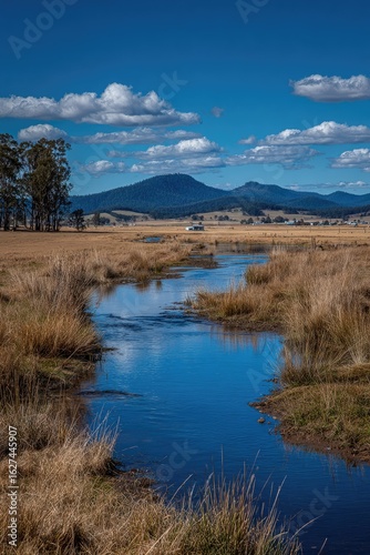Serene creek meanders through a tranquil landscape, reflecting a clear blue sky and distant mountains