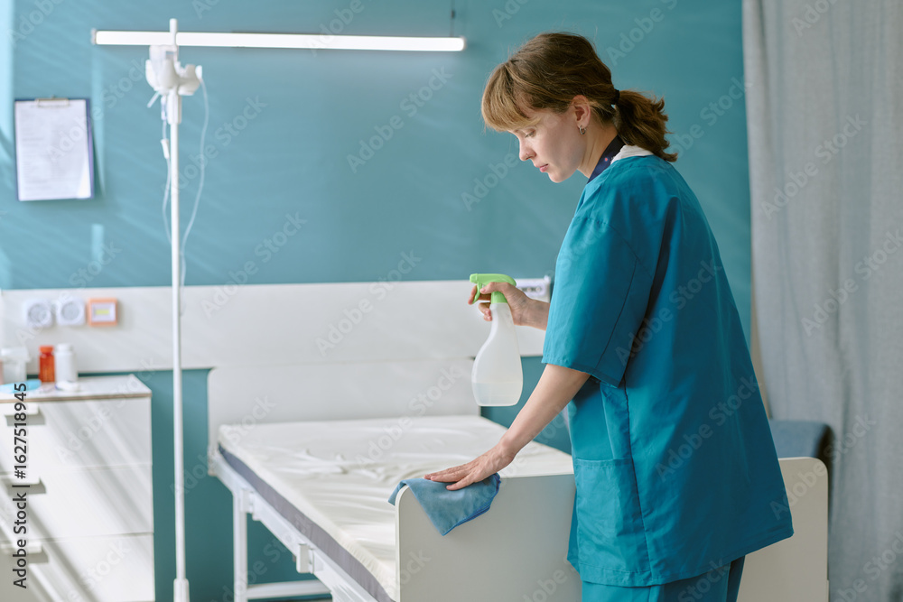 Fototapeta premium Caucasian young adult woman wearing medical scrubs cleaning hospital bed with spray bottle and cloth, standing in patient room, focusing on disinfecting surface for hygiene