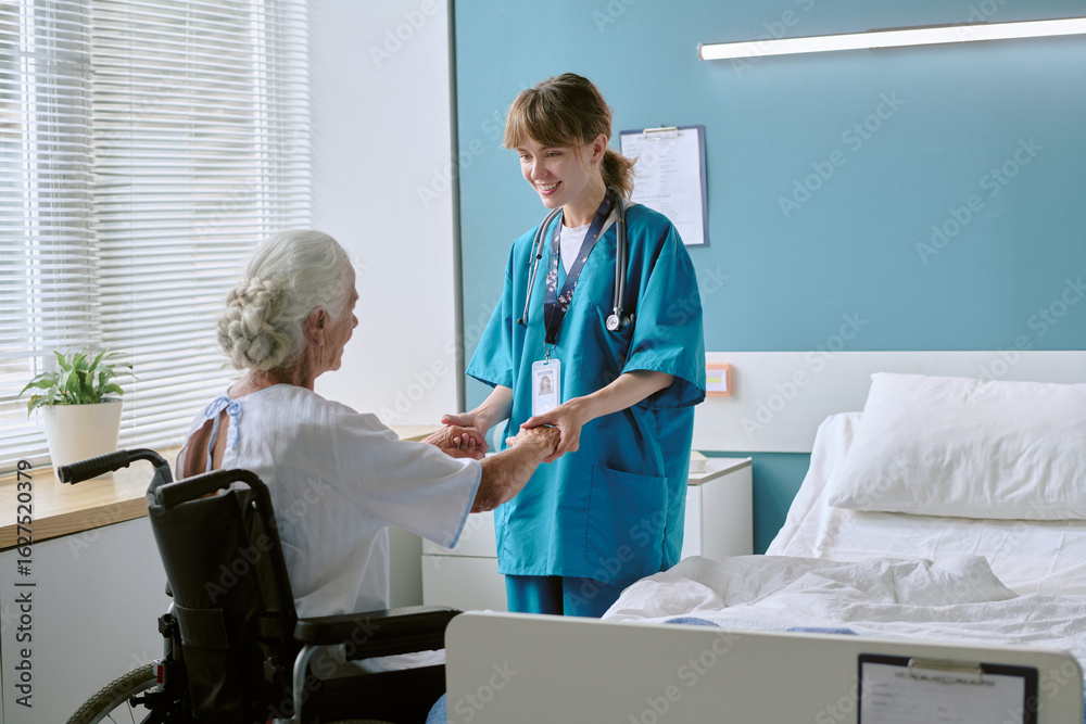 Obraz premium Caucasian senior woman with disability sitting in wheelchair holding hands with young adult Caucasian female nurse smiling in hospital room during medical care interaction