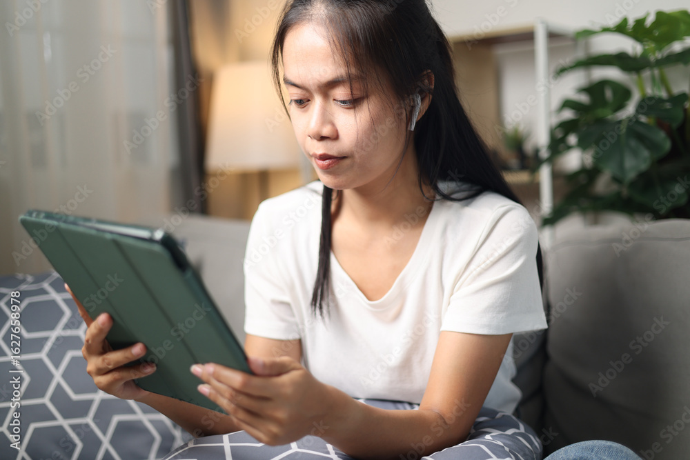 Fototapeta premium Portrait of Young Asian woman uses a tablet in relaxation while listening to music through wireless headphones on a sofa in a living room at home.