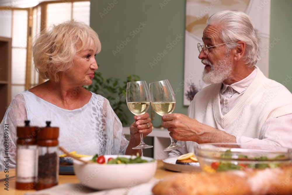 Elderly couple clinking glasses of white wine during dinner at table indoors Elderly couple clinking glasses of white wine during dinner at table indoors