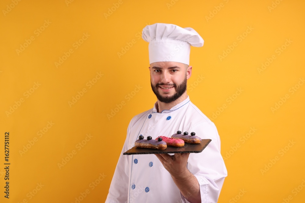 Happy confectioner in uniform holding eclairs on orange background Happy confectioner in uniform holding eclairs on orange background
