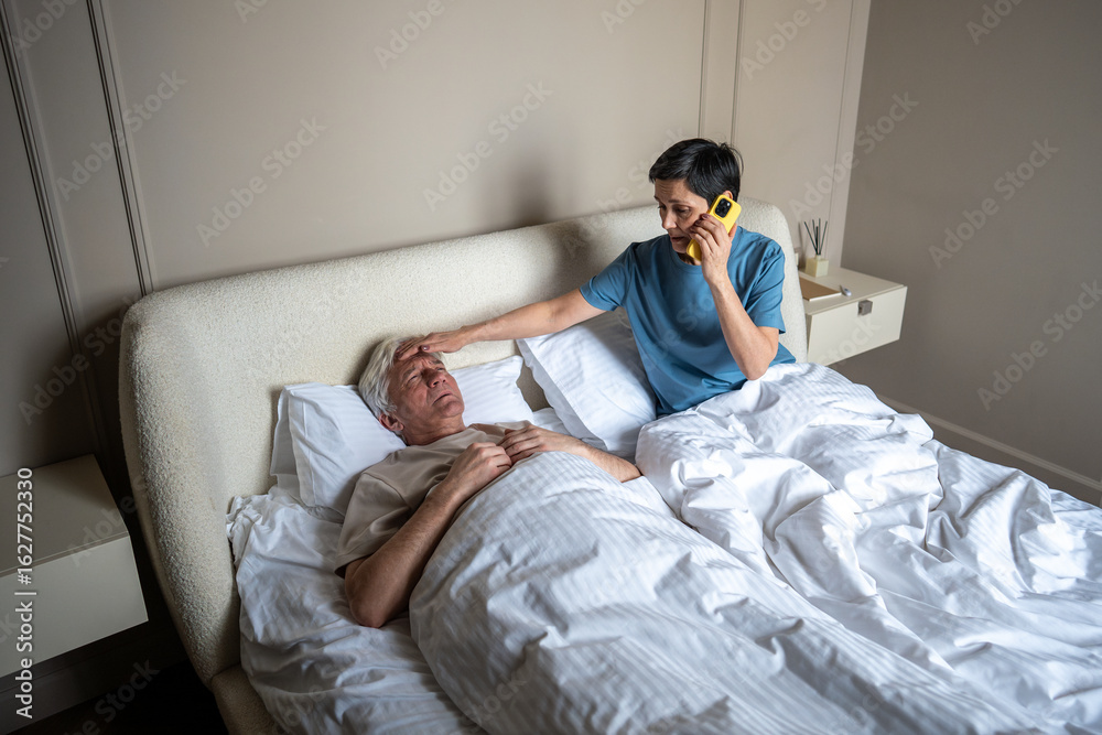 Obraz premium High angle view of worried wife touching feverish forehead of husband suffering from flu in bed, calling to inform his workplace. Illness care, family responsibility, health concern, domestic nursing