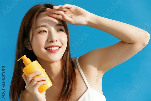 A young Asian woman smiling and shielding her eyes from the sun while holding a bottle of sunscreen.
