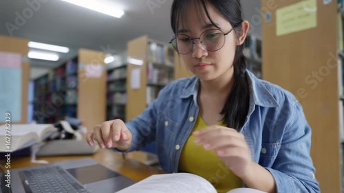 A focused young Asian female student with glasses uses a laptop and books to do research in a library, representing digital learning, hybrid education, and student lifestyle concepts.