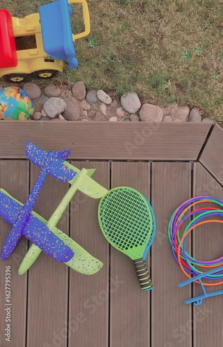 Backyard photo with a pile of outdoor toys for kids. A bunch of sports toys are lying on a wooden patio. Tennis rackets, plastic hoops, planes made of foam, and toy vehicle are beloved toys for boys. 