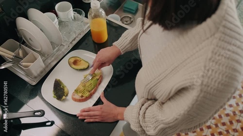 A woman spreads avocado on toast in the kitchen, orange juice in the background. She is making a healthy, nutritious breakfast.